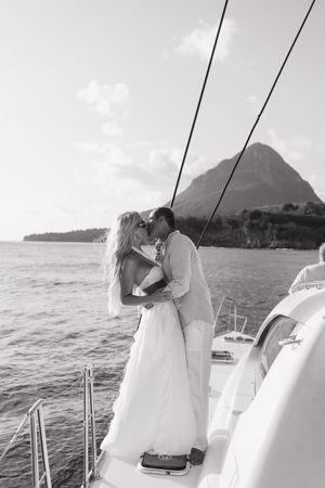 Black and white photo of a couple kissing on a sailboat with a tropical mountain backdrop on their wedding day.