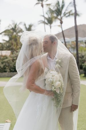 Bride and groom sharing a romantic kiss under flowing wedding veil with tropical palm trees backdrop.
