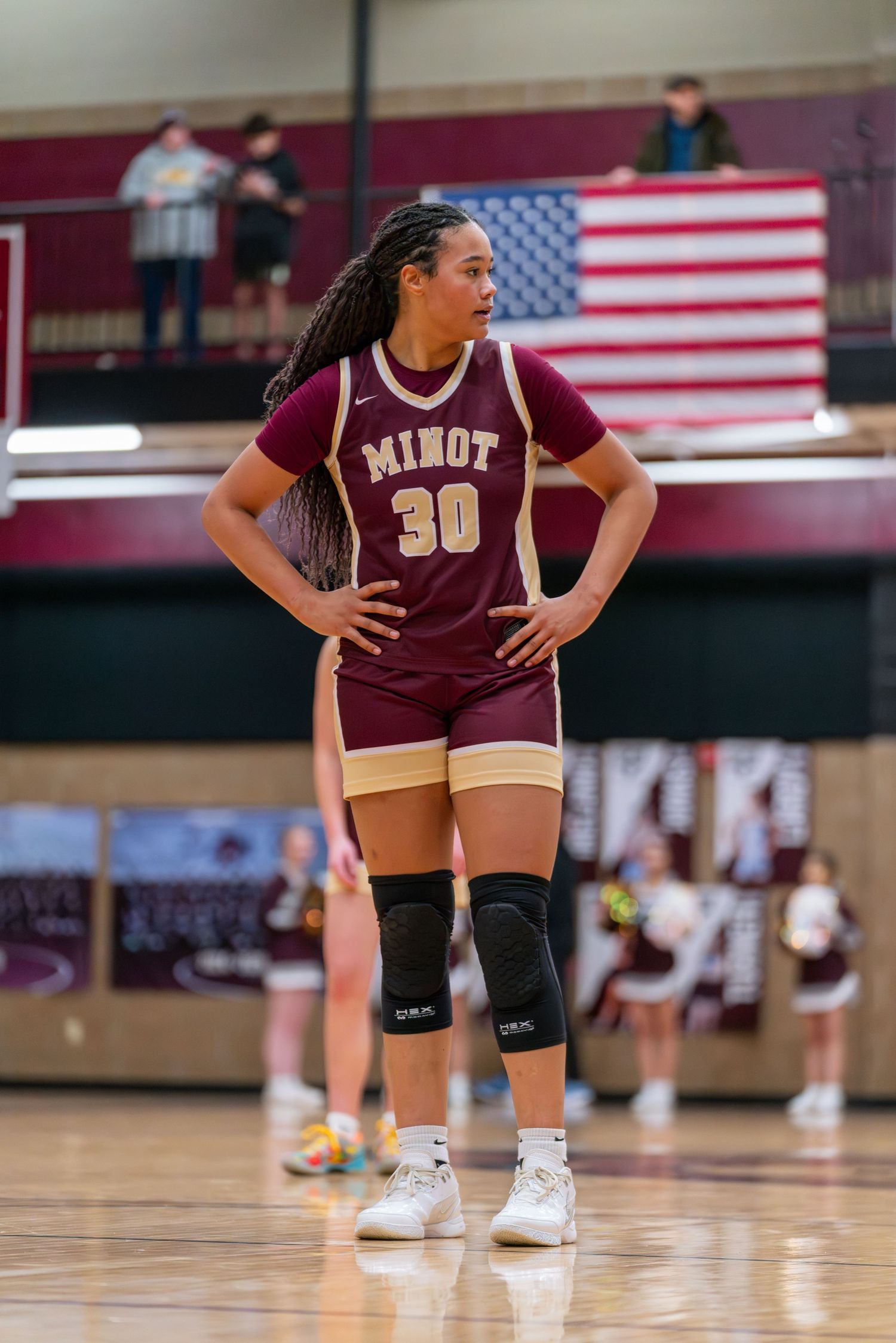 Basketball player wearing jersey number 30 and maroon Minot uniform stands on court with American flag in background.