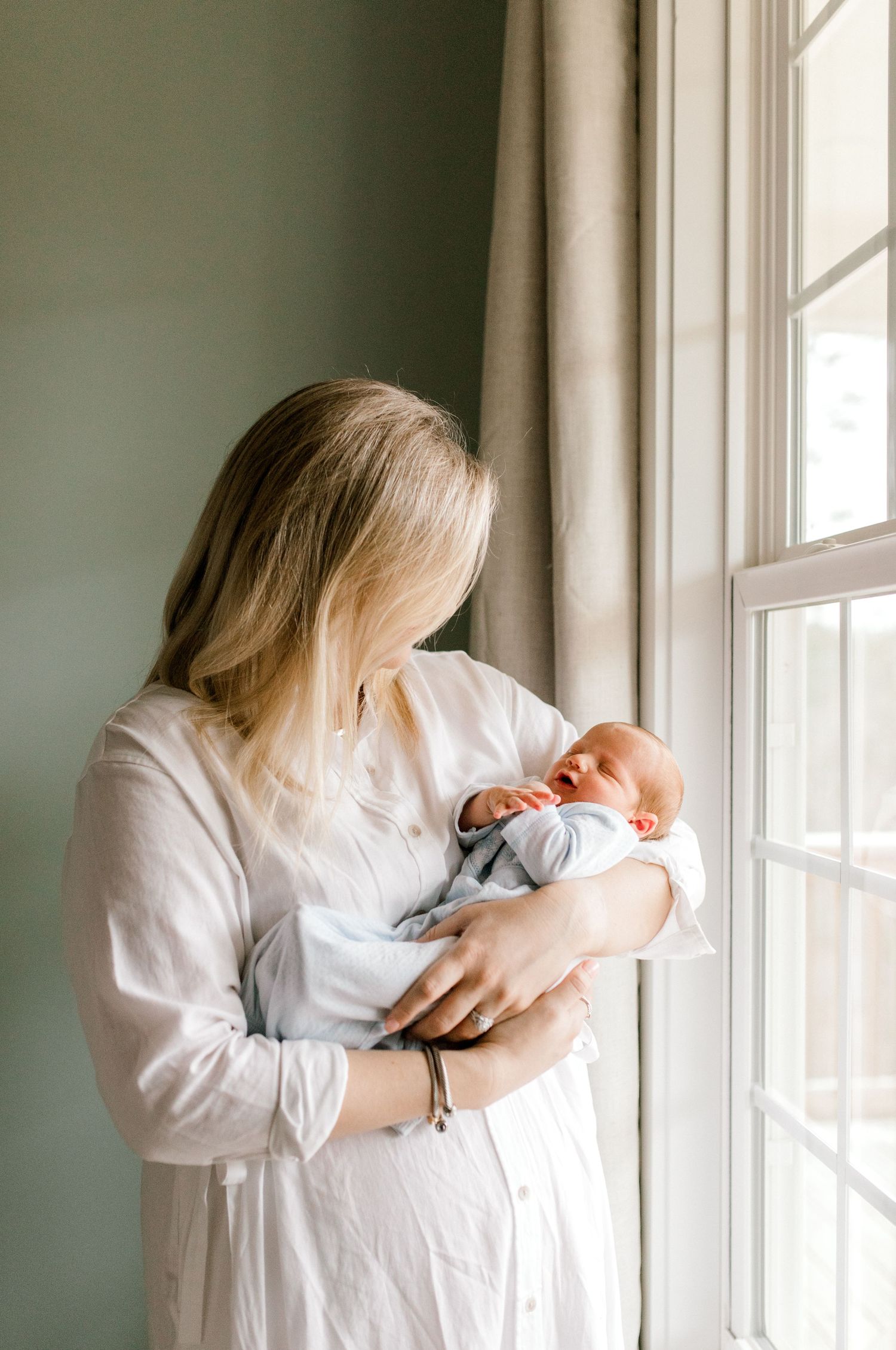 Mother in white robe holds newborn baby while standing by window in natural light.