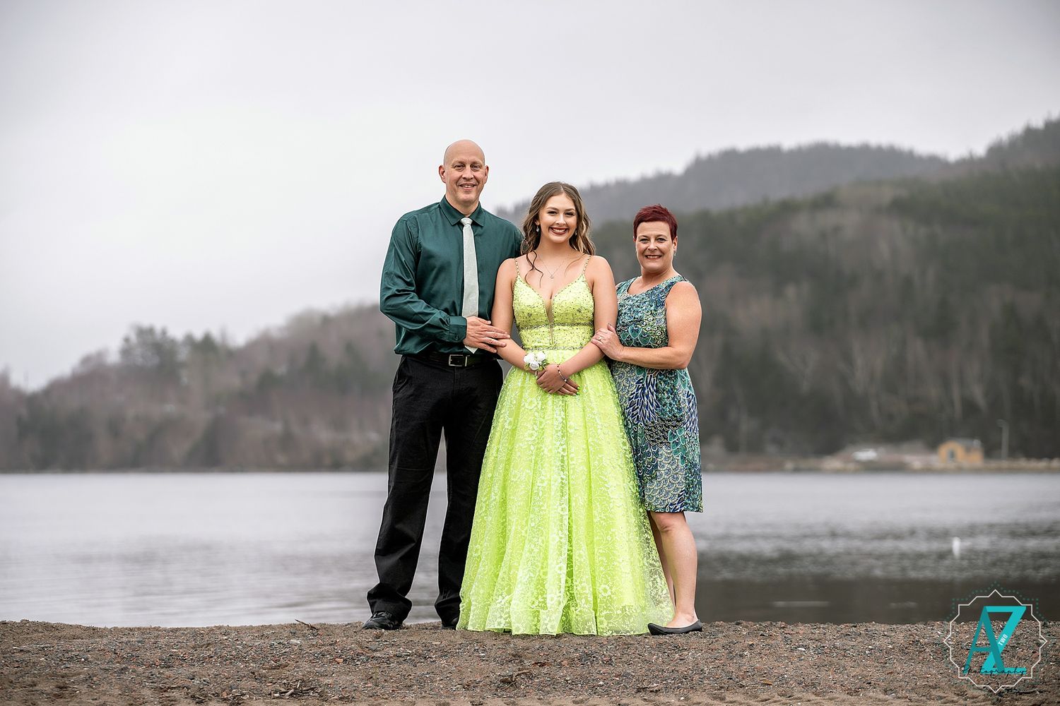 Three people pose together on beach with mountains in background, center figure in lime green gown.