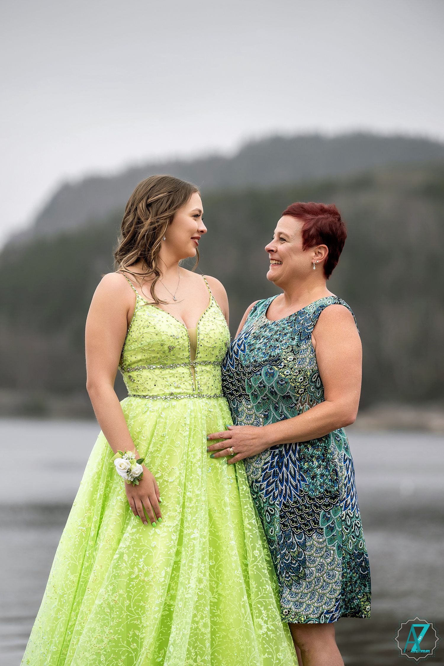 Two people share a warm moment by the water, wearing formal dresses in lime green and blue patterns.