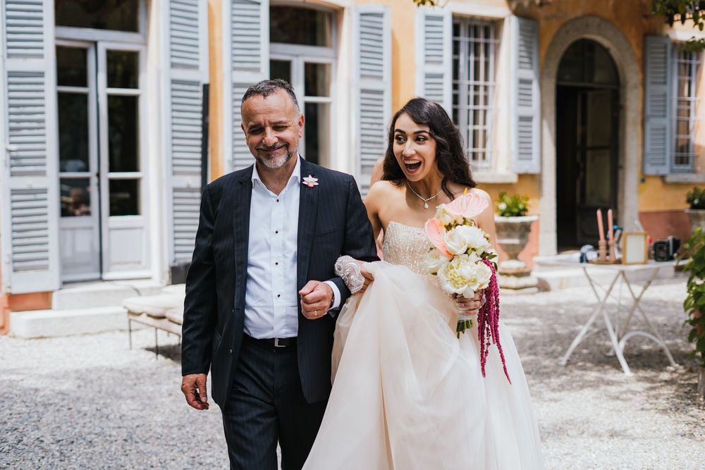 Laughing couple walks arm in arm outside a charming French country house with blue shutters.