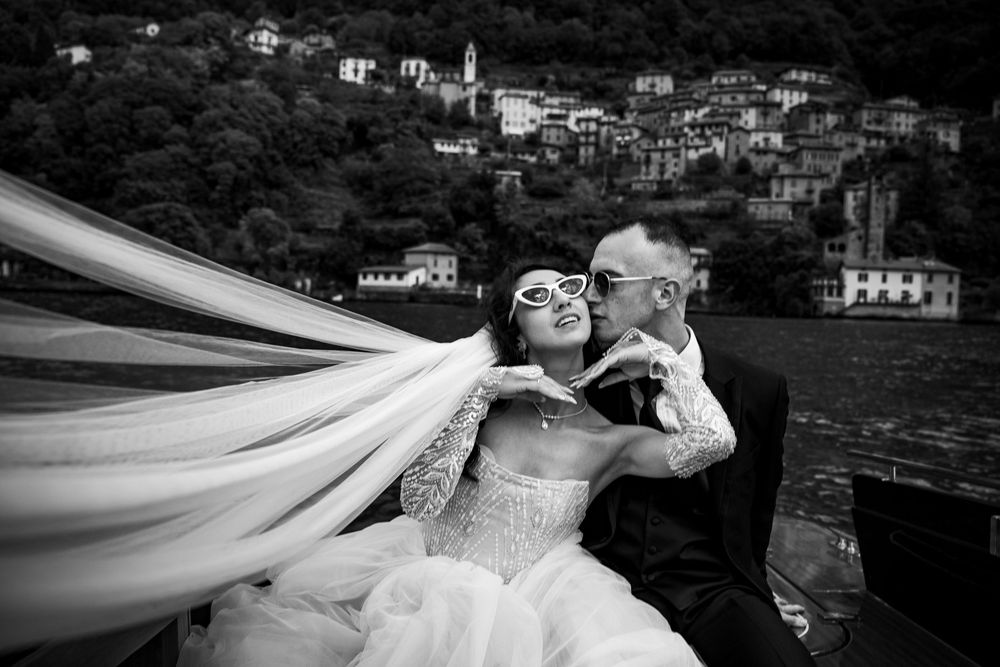 A romantic wedding photo in black and white with flowing veil captured by the lake, with hillside village in background.