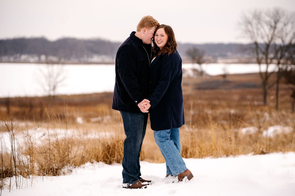 A couple in winter coats embraces while standing in a snowy field with tall grass and a lake in the background.