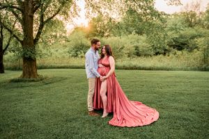 A couple poses in a grassy field, with her wearing a flowing coral dress during golden hour.