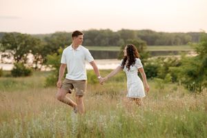 Couple holding hands walking through a sunny green meadow near a lake at golden hour sunset.