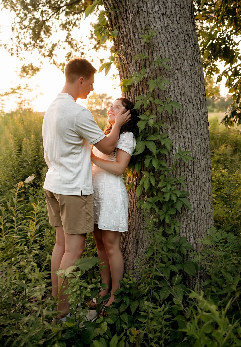 A couple shares a romantic moment by a tree in a sunlit meadow, the woman in a white dress smiling up at her partner.