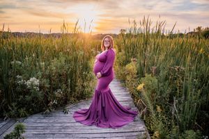 A woman in a flowing purple maternity dress poses on a wooden boardwalk at sunset.