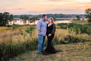 A couple stands together in a grassy field overlooking a lake at sunset.