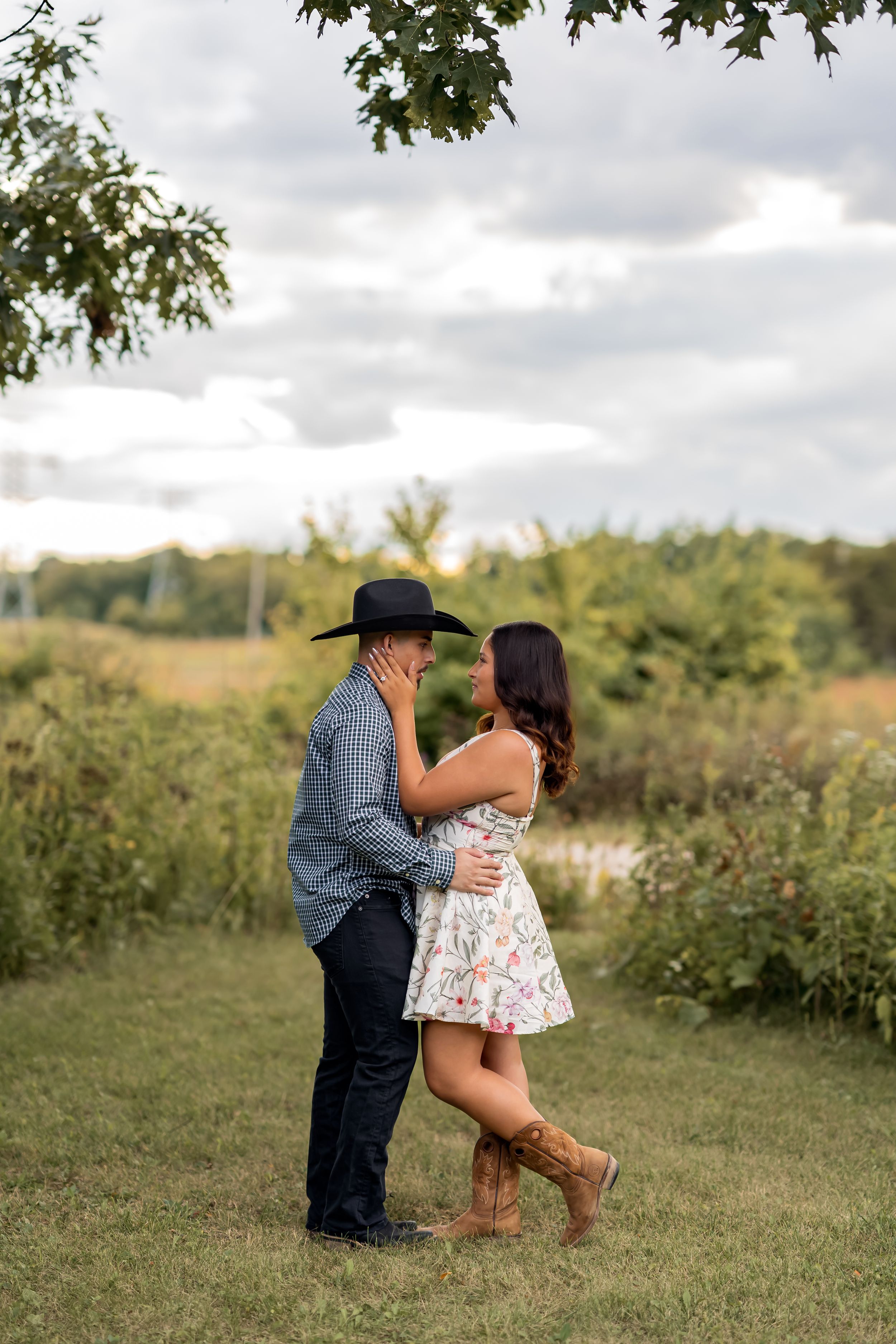 Couple embracing in a lush green field, man in cowboy hat and plaid shirt, woman in floral dress and cowboy boots.