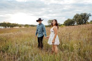 Couple holding hands in golden field, man in cowboy hat and blue shirt, woman in white dress, cloudy sky.