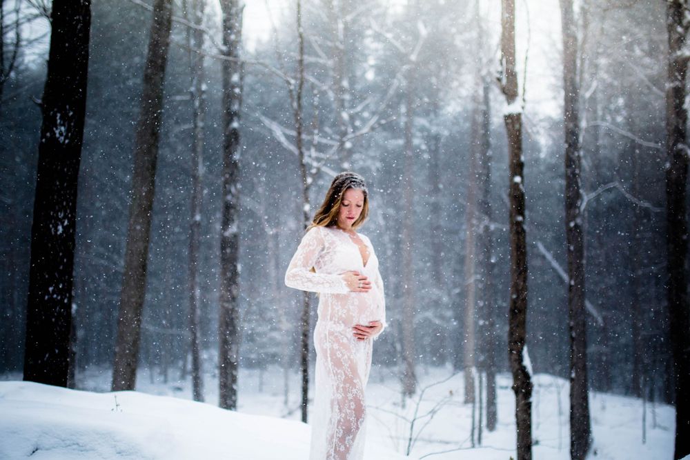 A dreamy maternity photo featuring a lace dress against a snowy forest backdrop.
