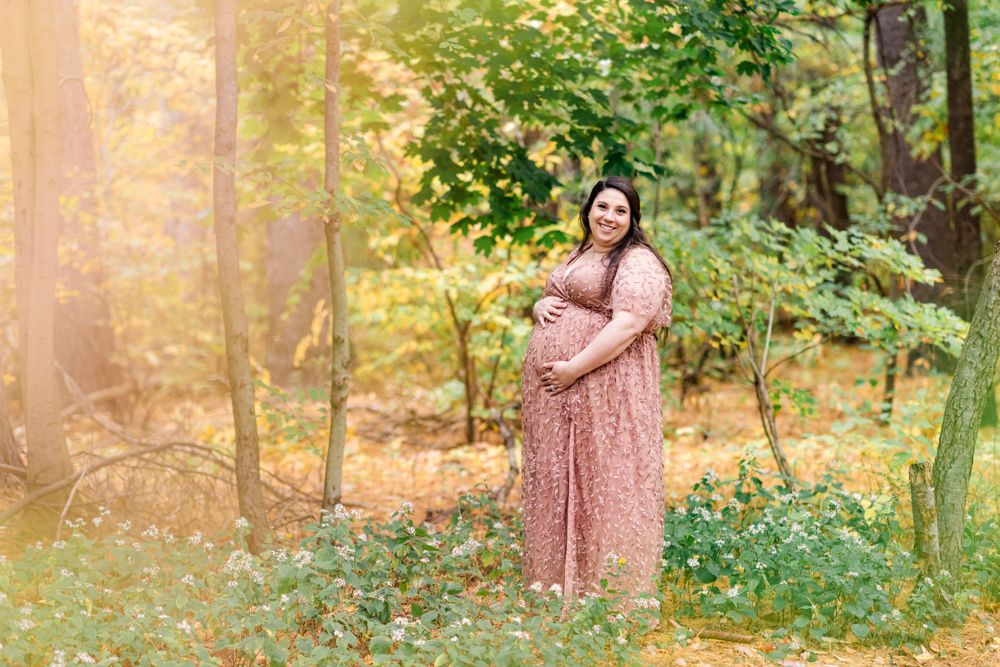 A maternity portrait in a pink lace dress surrounded by autumn foliage in golden sunlight.