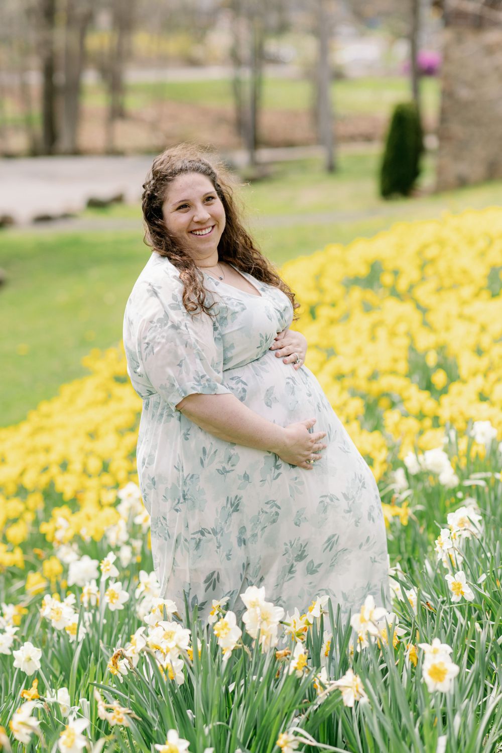 Expectant mother in white dress surrounded by blooming spring daffodils and trees.