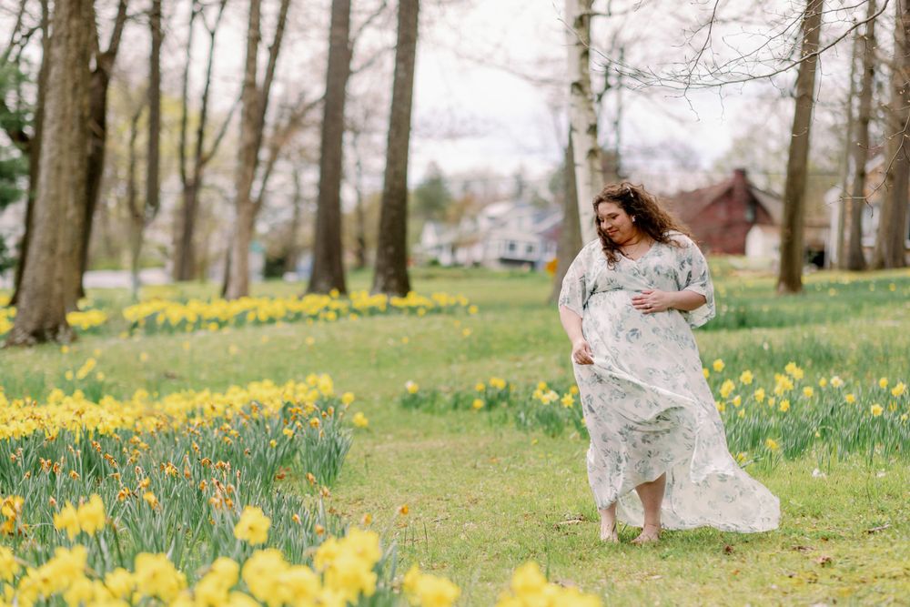 Spring maternity photoshoot in a field of blooming yellow daffodils with flowering trees in the background.