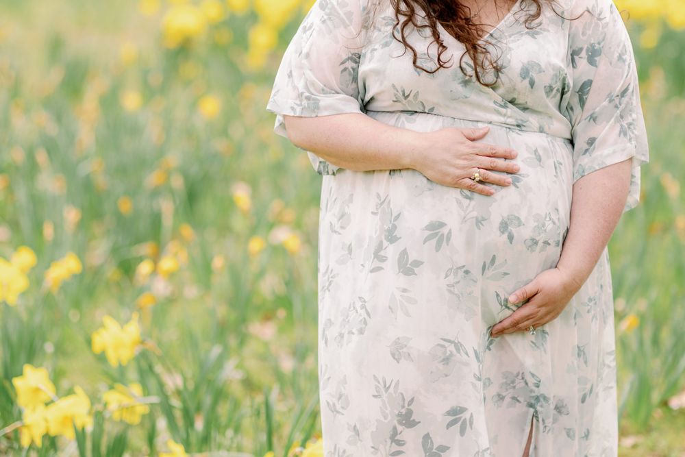 Woman in floral dress poses among yellow daffodils during spring maternity session.