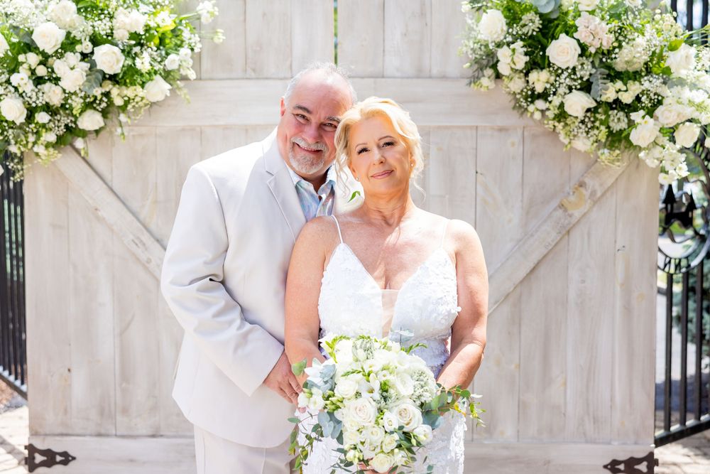 Wedding couple embraces and smiles in front of white stone wall with elegant floral decorations.