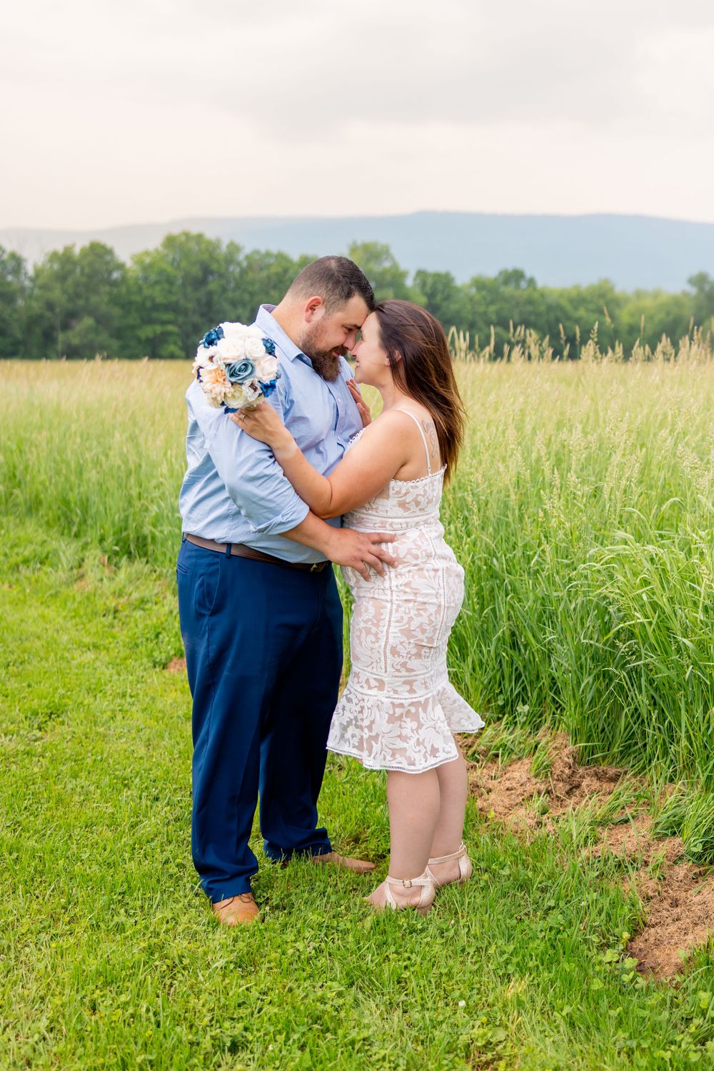 A couple dances together in a scenic green meadow with mountains visible in the background during a summer day.