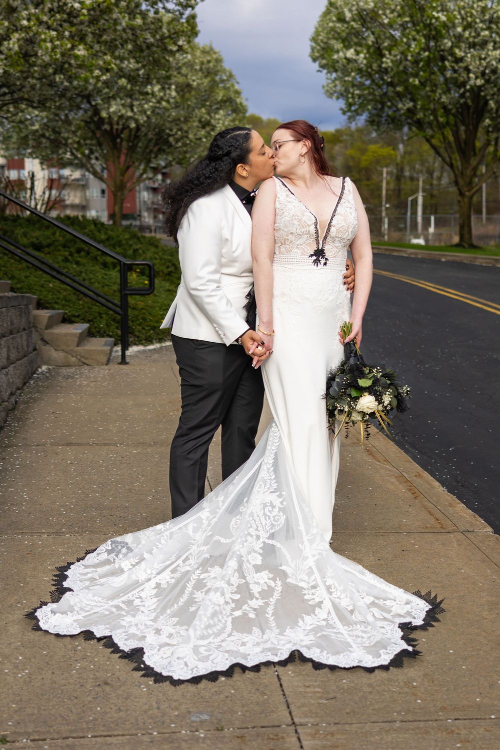 A romantic newlywed couple embraces on a tree-lined street in their wedding attire, with a flowing lace train trailing behind.