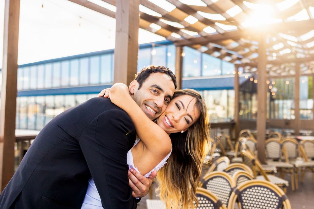 A joyful couple laughs together while sitting at a bistro table with woven chairs on a sunlit restaurant patio.
