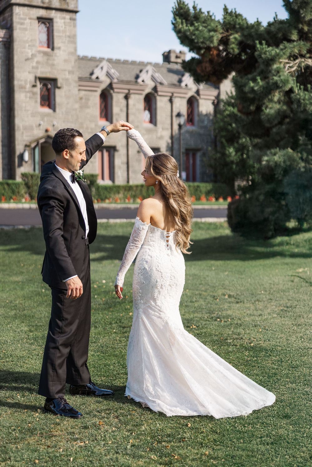 A couple in wedding attire dances playfully on a green lawn with a historic stone church building in the background.