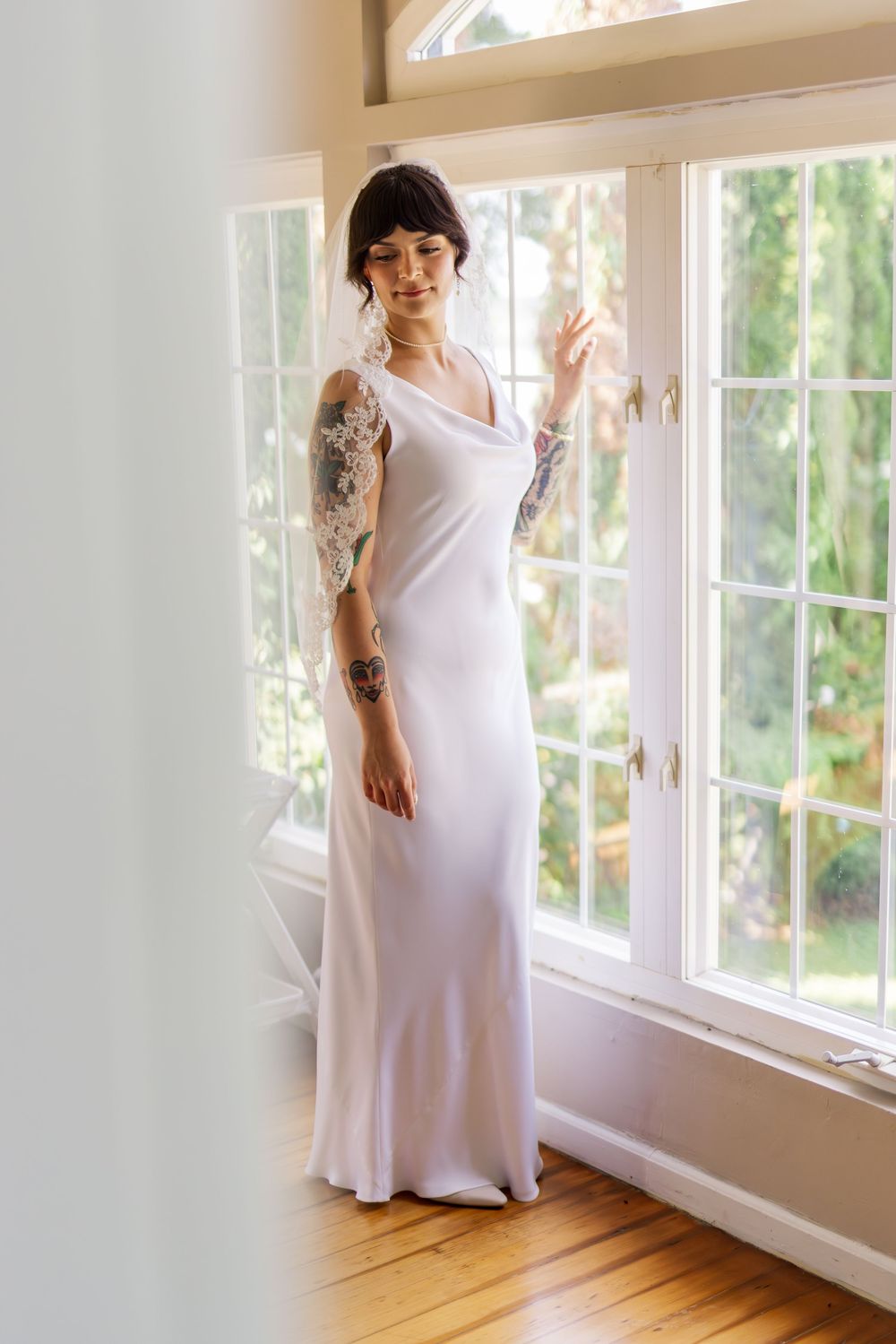A person in a flowing white maxi dress stands in front of sunlit windows with wooden floors and natural lighting.