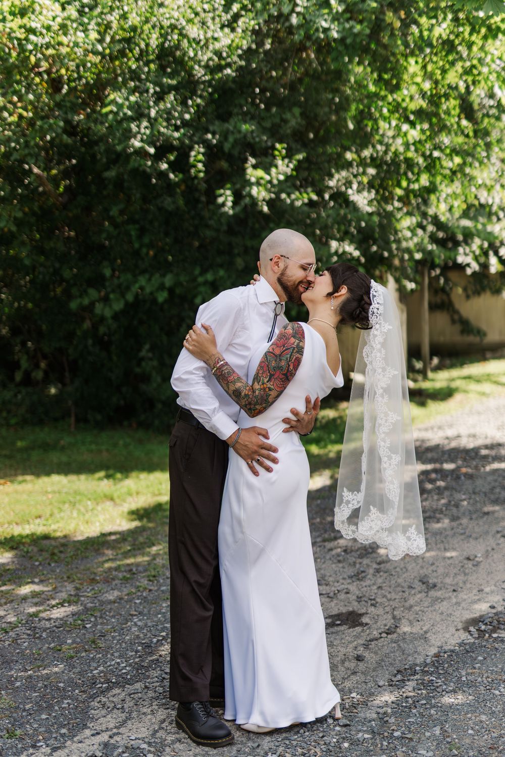 A romantic first look between newlyweds sharing a kiss outdoors with green trees in the background.