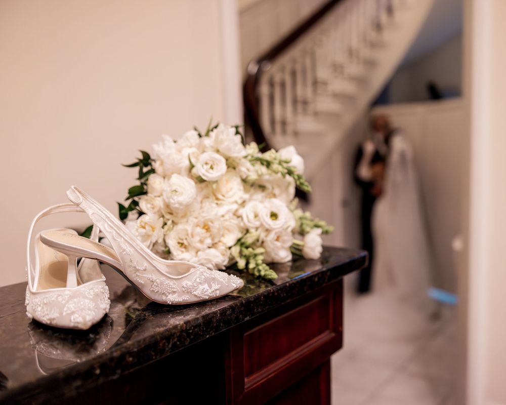 White wedding shoes and floral decorations arranged elegantly on a brown wooden surface in warm lighting.