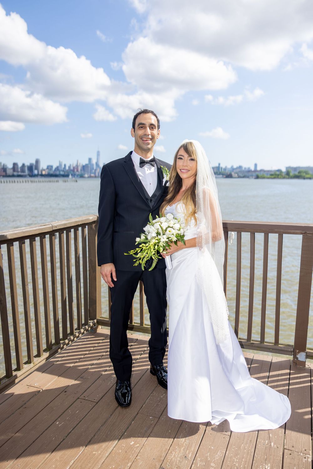Wedding couple poses on wooden deck overlooking city skyline and waterfront on sunny day.