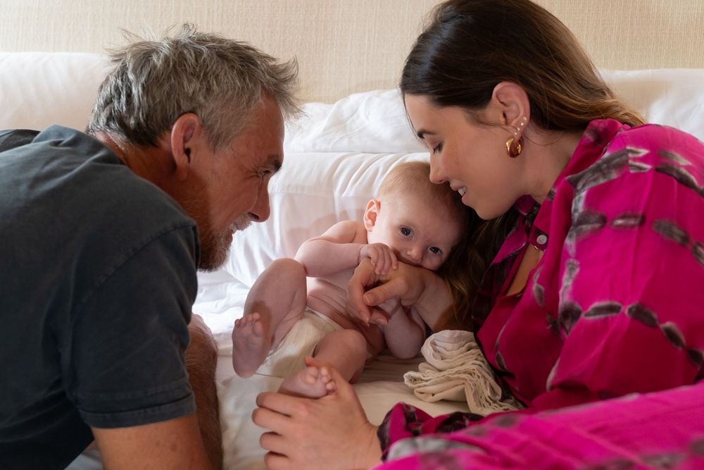 New parents cuddling their newborn baby during an in‑home first photo shoot, natural light lifestyle family photography by SRX Media Group i