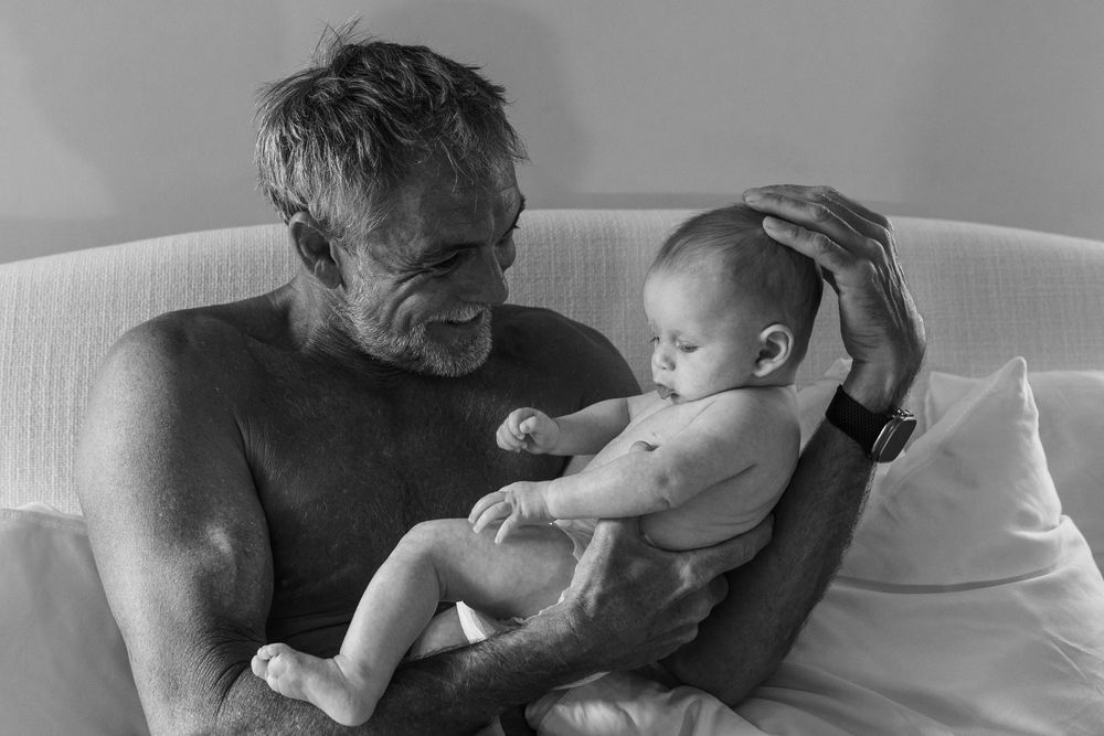 New parents cuddling their newborn baby during an in‑home first photo shoot, natural light lifestyle family photography by SRX Media Group i