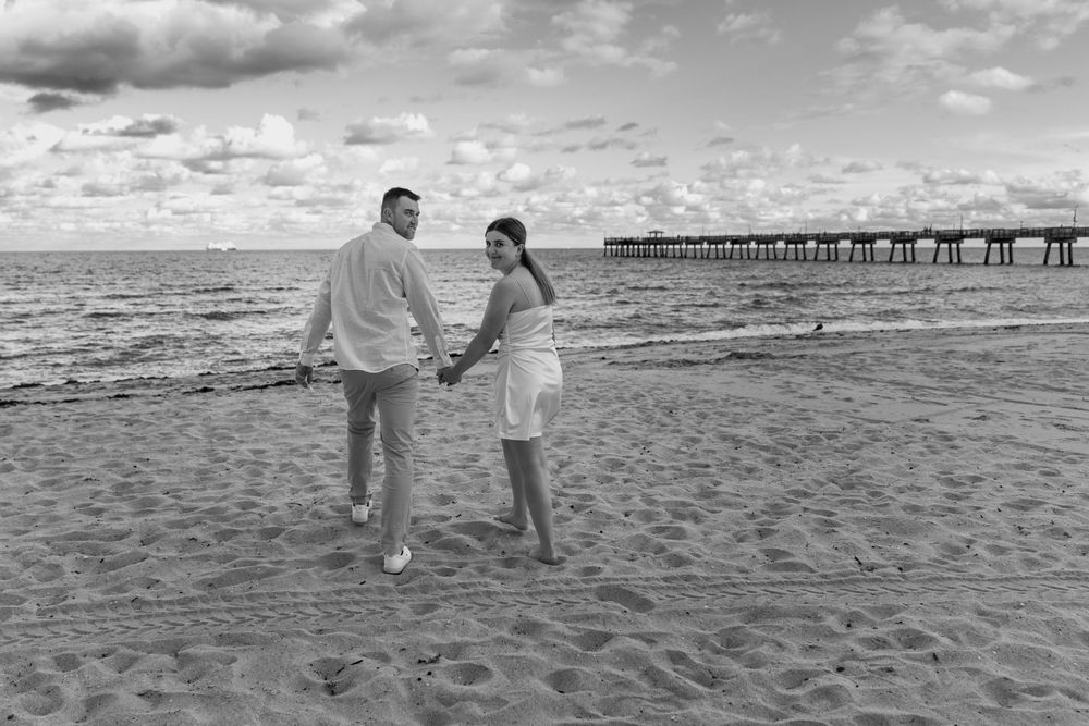 Married couple walking on a Fort Lauderdale beach at sunset, looking back with ocean background black and white