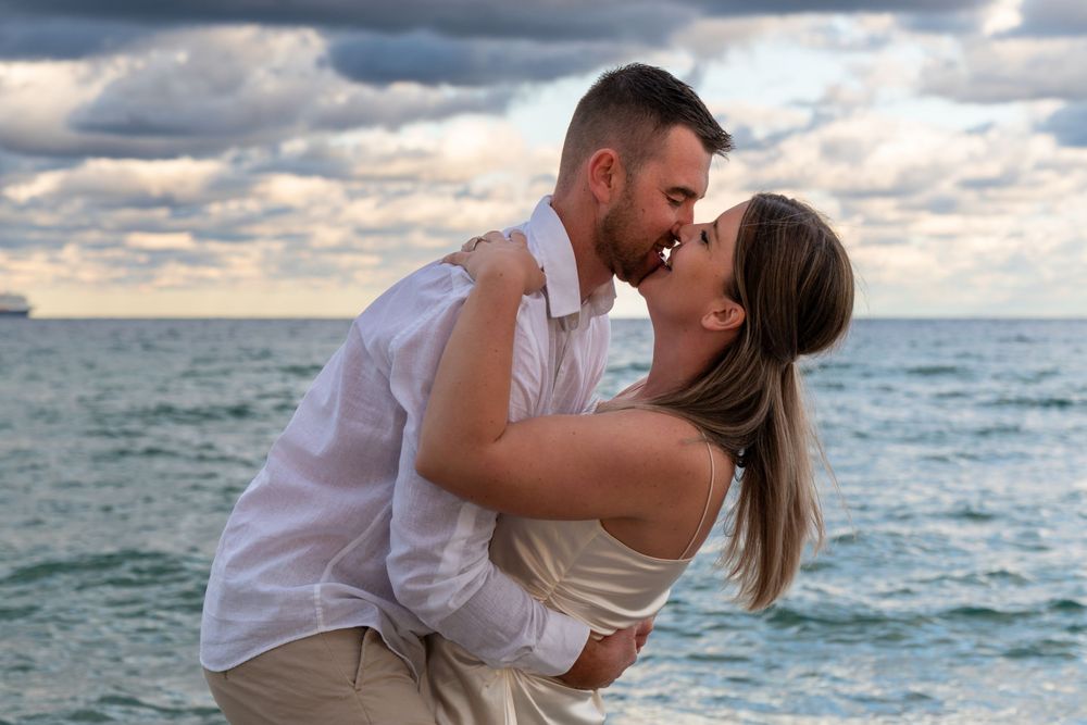 Married couple embracing on a Fort Lauderdale beach at sunset, looking at each other with ocean waves and dramatic clouds in the background