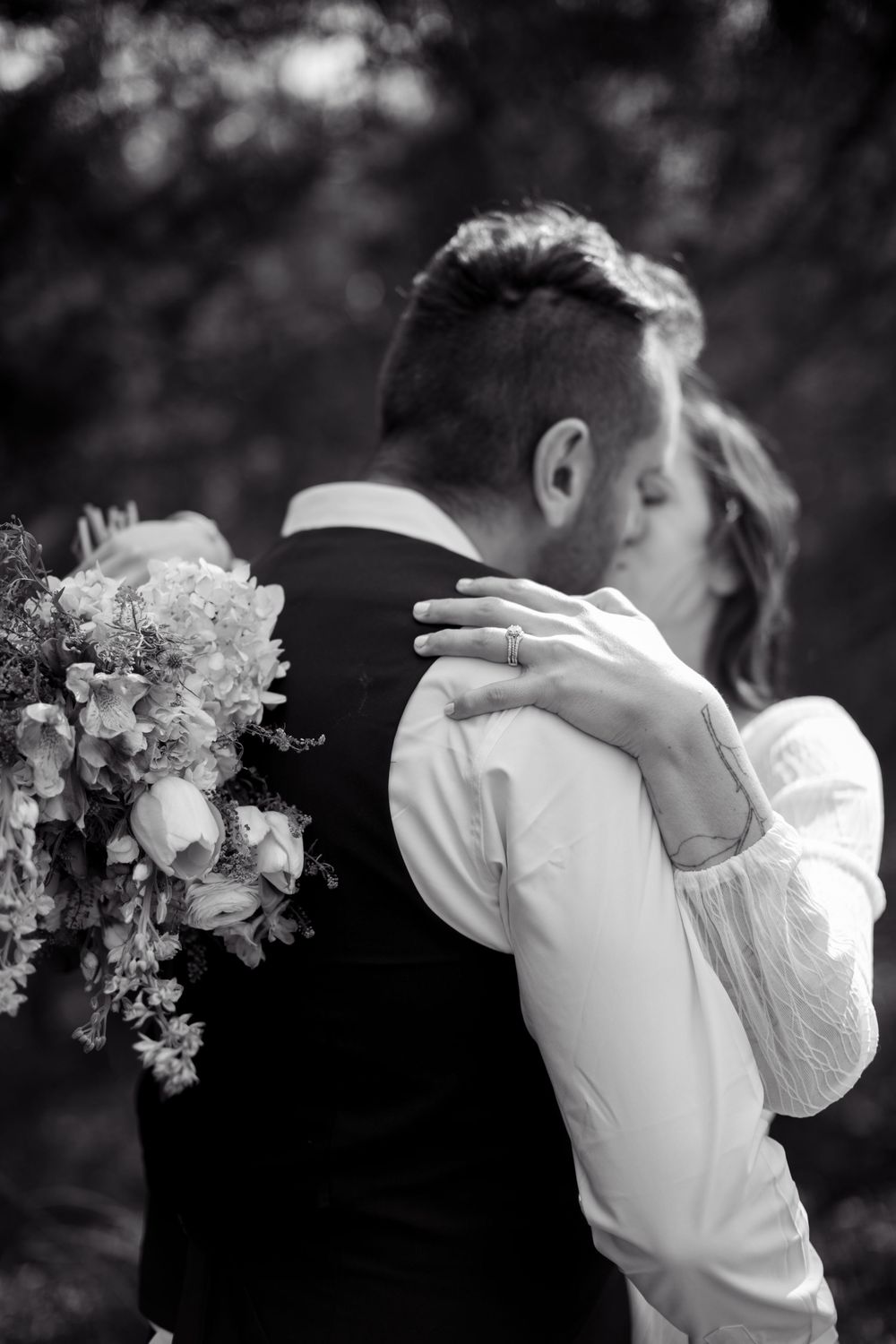 Black and white photo of a bride and groom sharing a kiss, bride holding a bouquet and resting her hand on grooms shoulder.