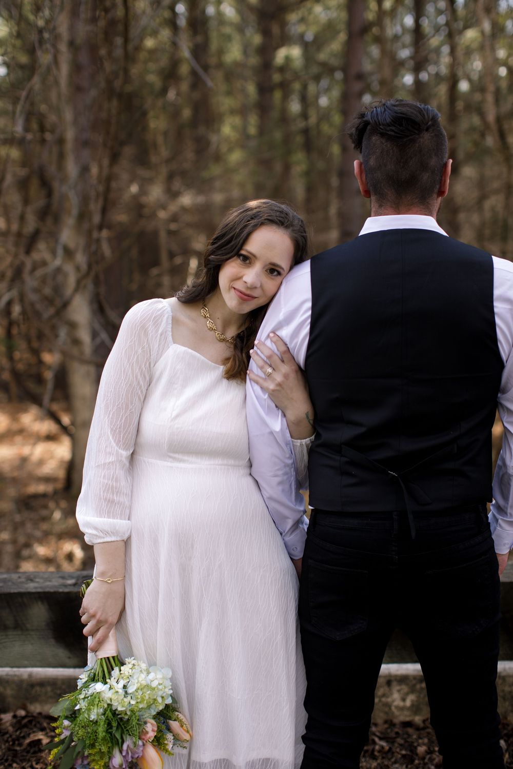 Pregnant bride in white dress leans on groom's shoulder holding wildflowers in a forest wedding setting.