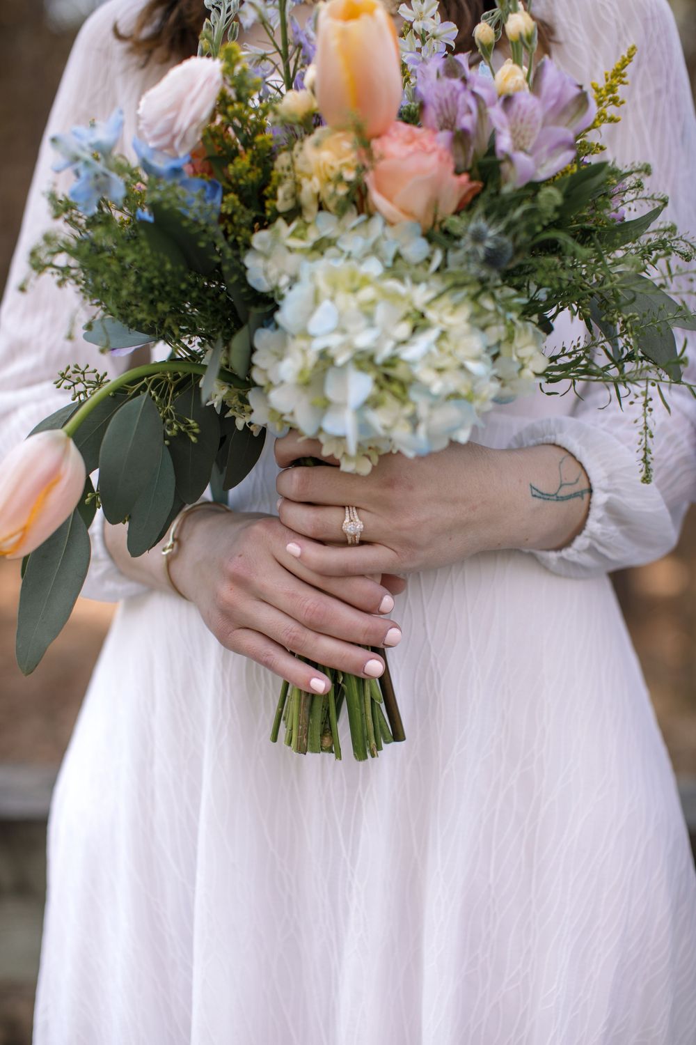 Bride in white dress holding colorful wedding bouquet with hydrangeas, roses, and greenery, wearing diamond engagement ring.