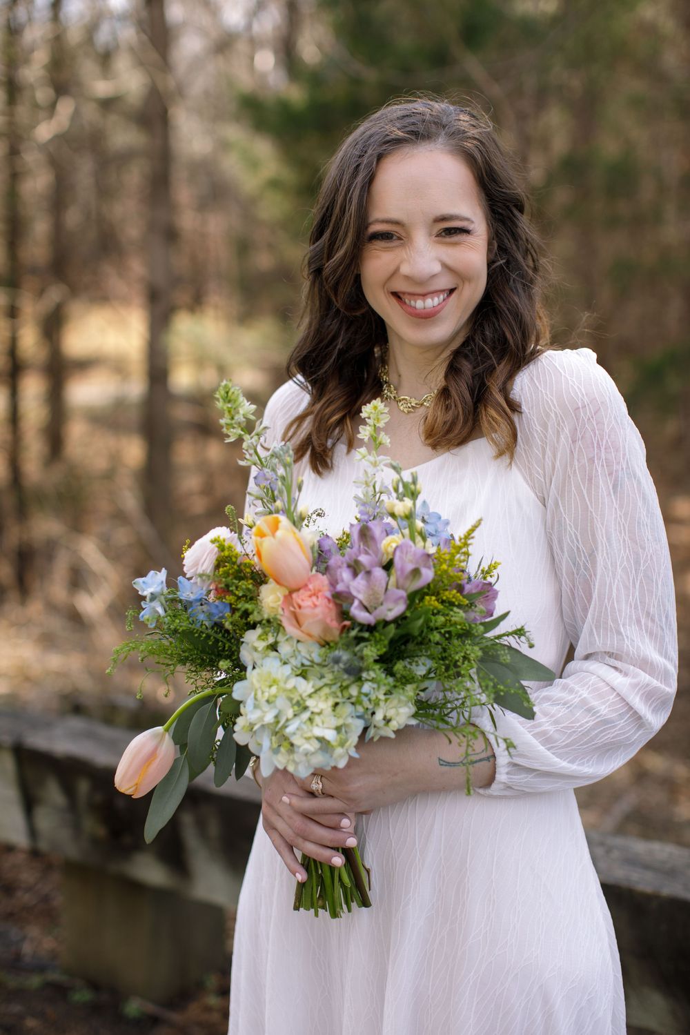 Smiling bride in white dress holding colorful spring bouquet with tulips and hydrangeas outdoors.