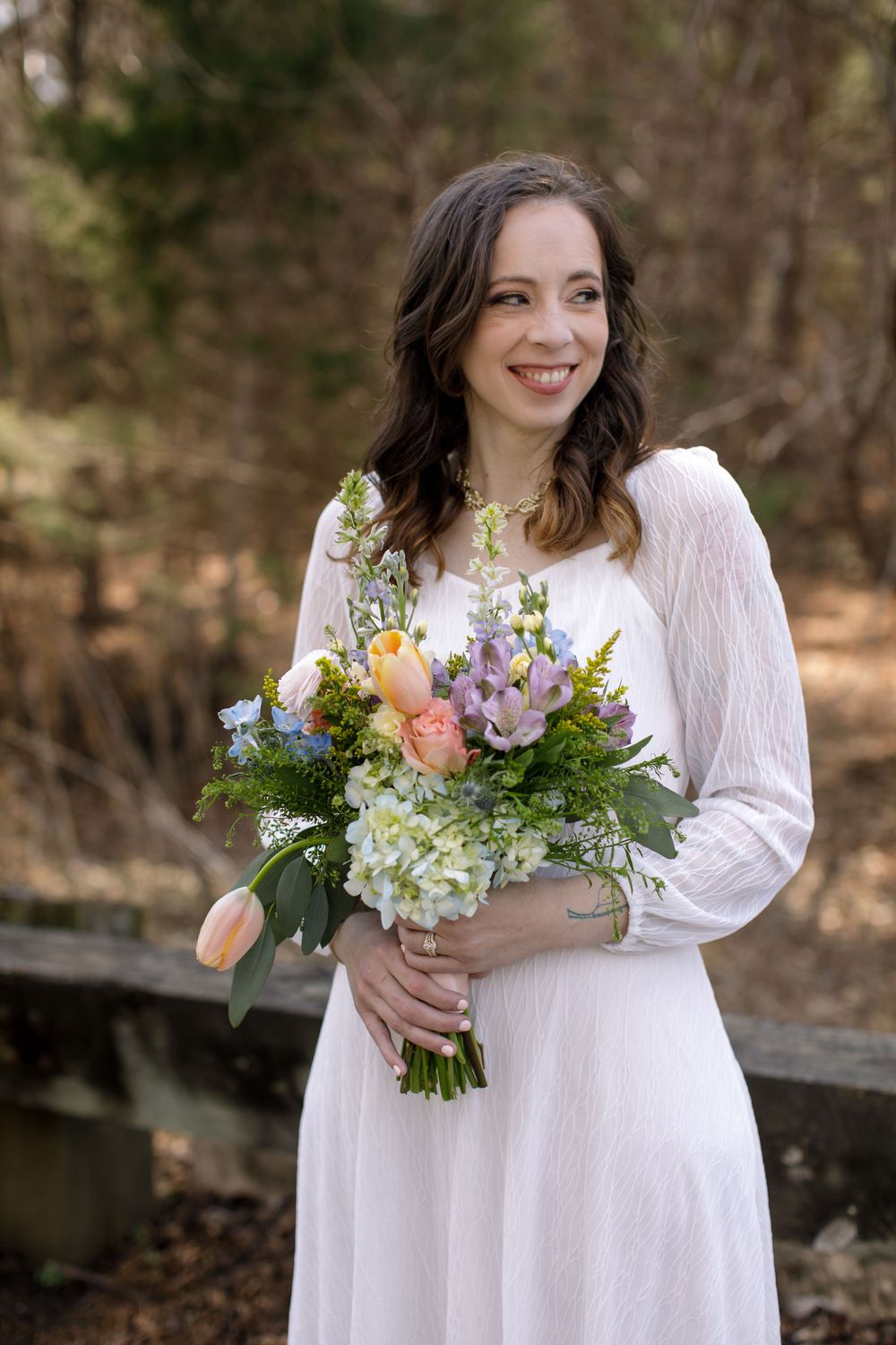 Smiling bride in white dress holding colorful spring bouquet with tulips and hydrangeas outdoors.