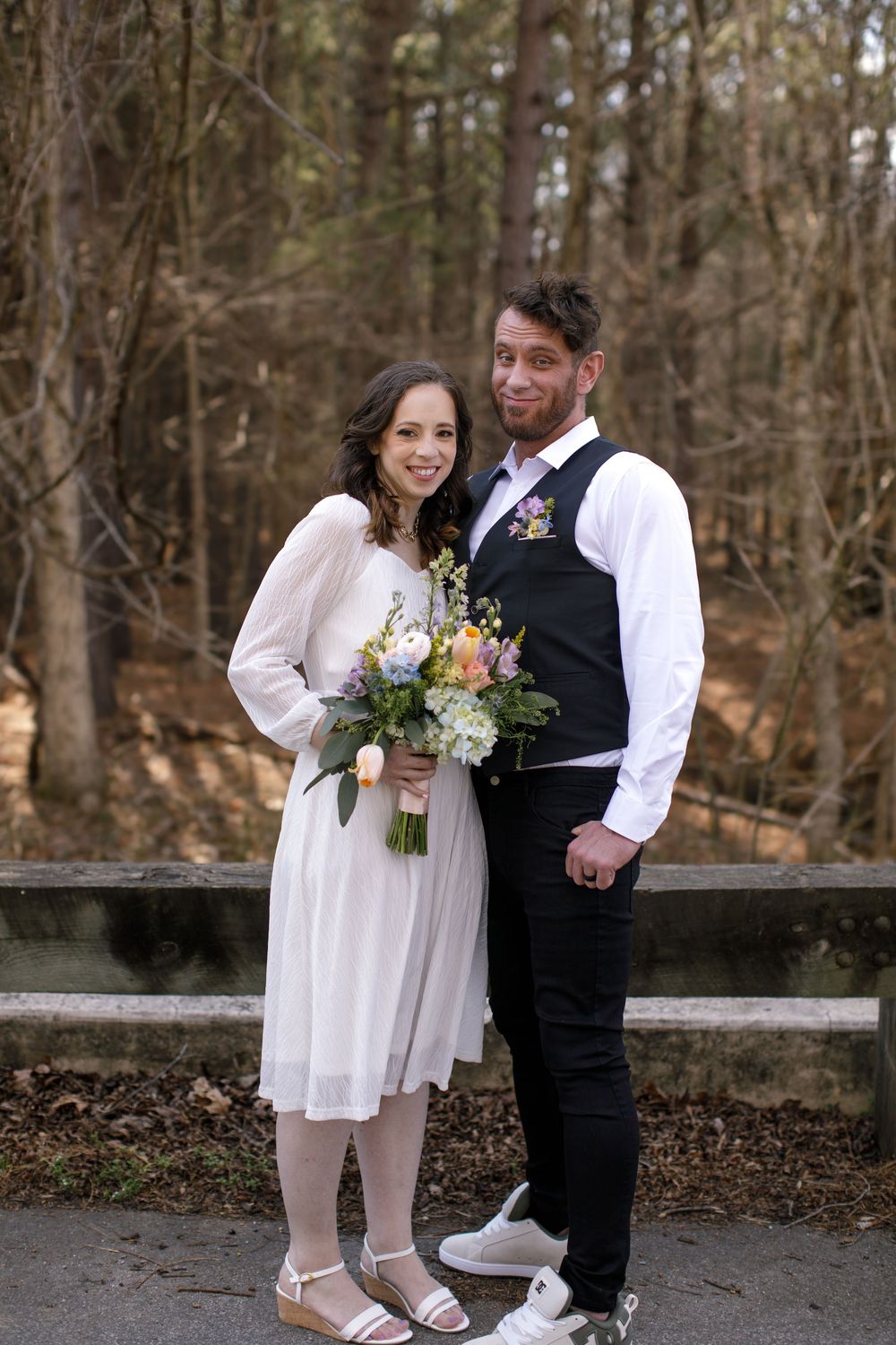 Bride and groom pose outdoors in forest setting, she holds colorful bouquet in white dress, he wears black vest.
