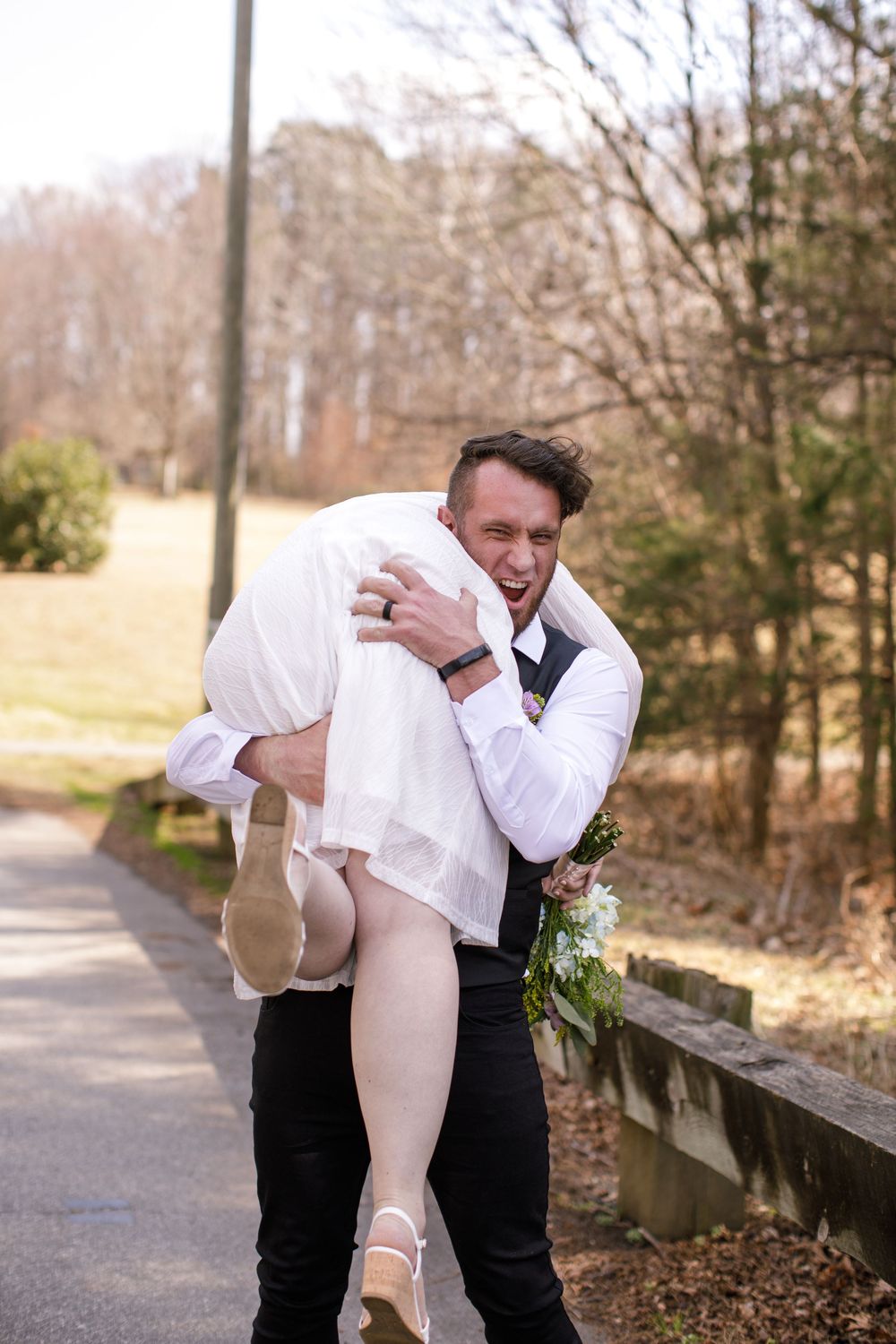 Groom carries bride over his shoulder laughing outdoors on a path, wearing wedding attire with bouquet.