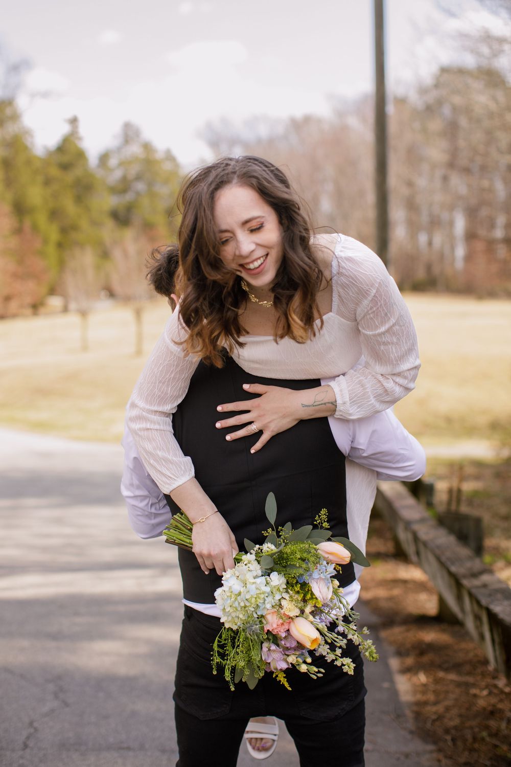 Laughing woman in white blouse holds spring bouquet while being hugged from behind outdoors.