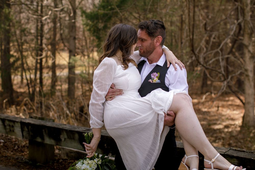 Groom carrying bride in white dress outdoors on wooden bridge surrounded by trees in romantic wedding photo.