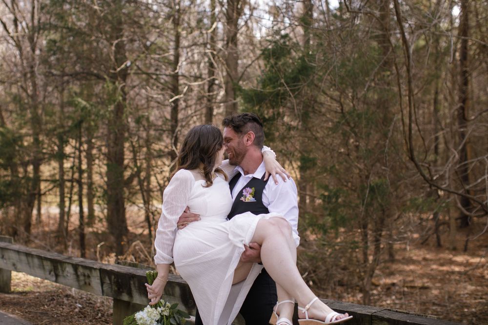 A couple shares a romantic moment on a wooden fence surrounded by bare trees, with the bride in a white dress.