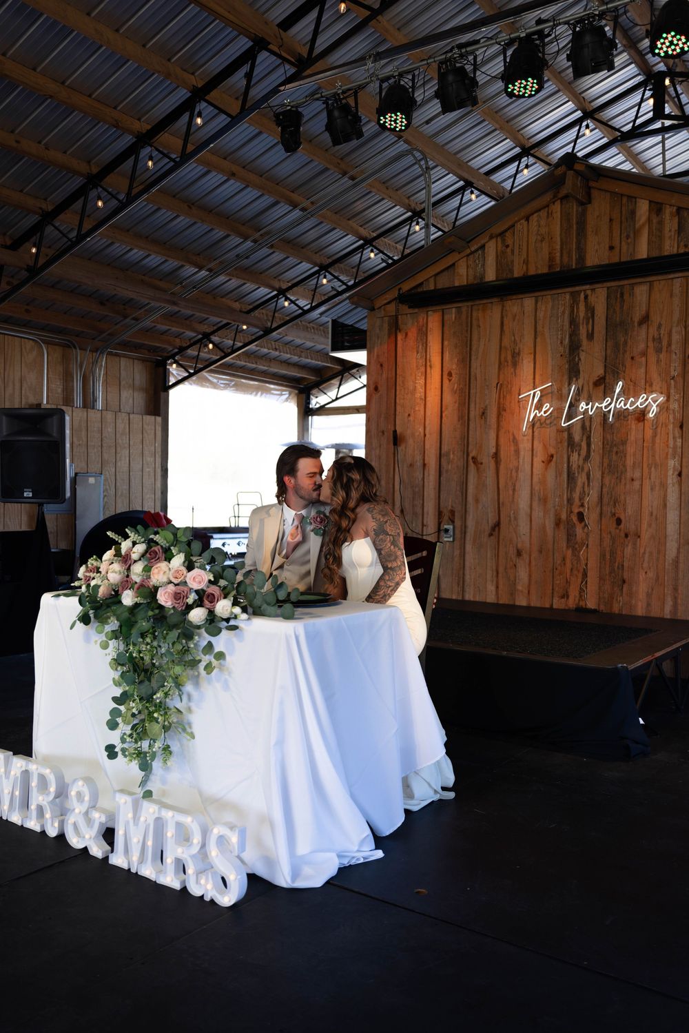 Newlyweds share a kiss at their sweetheart table decorated with MR & MRS letters and floral arrangements at a rustic barn venue.