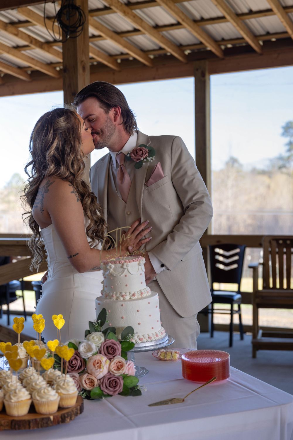 Bride and groom sharing a kiss at wedding cake table with cupcakes and floral decorations on rustic venue porch.