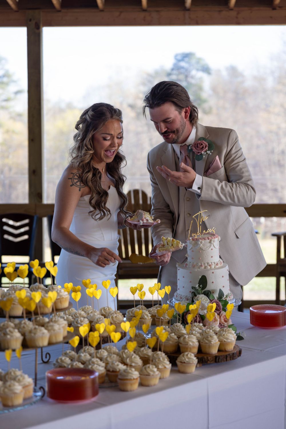 Bride and groom smiling while cutting their wedding cake surrounded by cupcakes with yellow flower toppers at rustic venue.