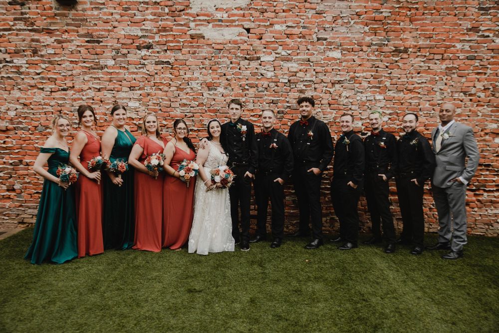 A wedding party poses against a brick wall with bridesmaids in colorful dresses and groomsmen in dark suits.