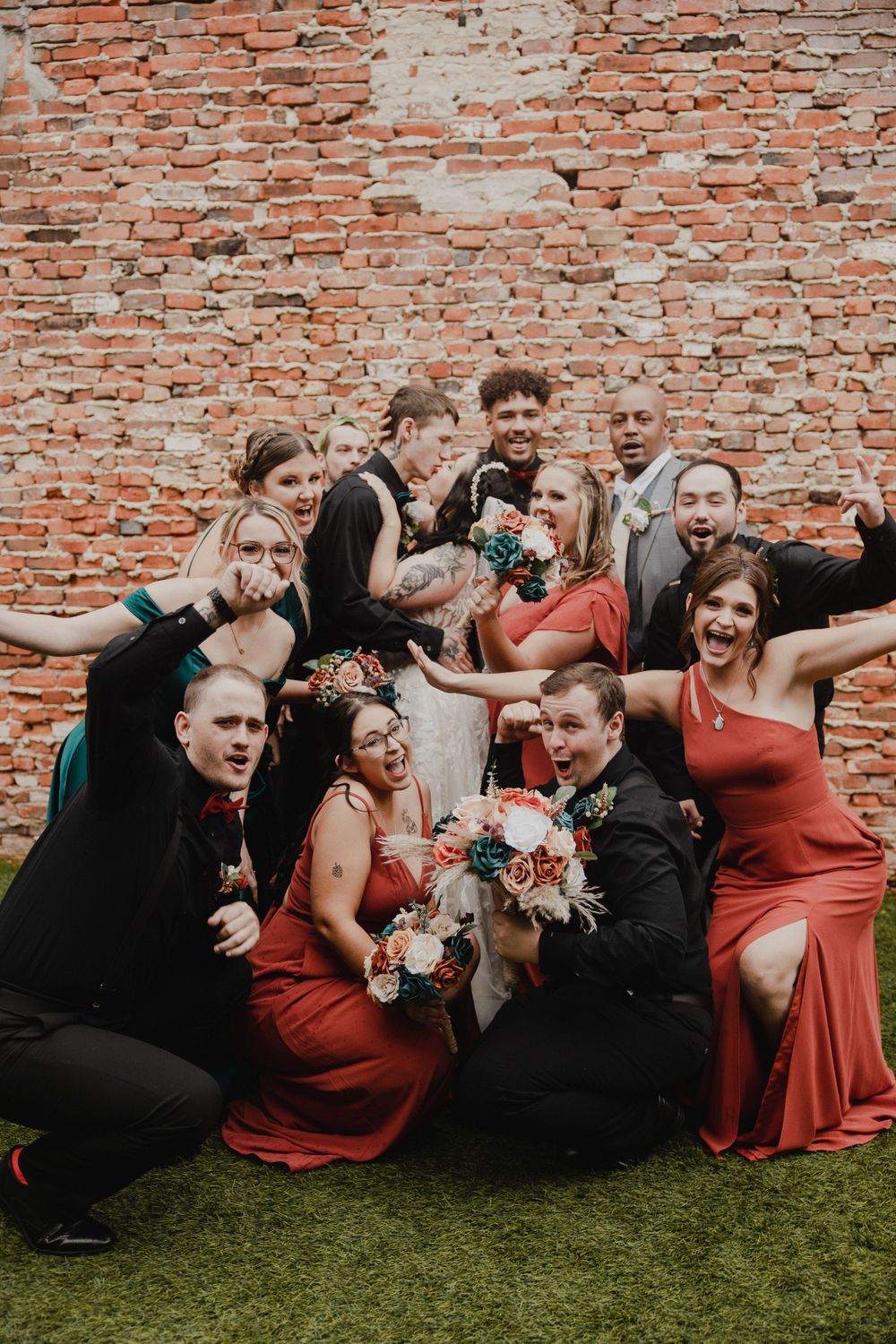 Wedding party in red dresses and black suits poses playfully against brick wall.