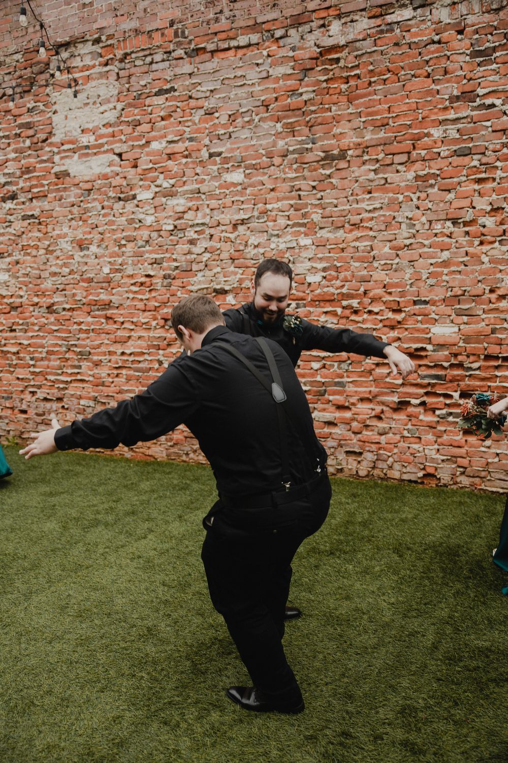 Two people in black suits playfully pose against a rustic brick wall with artificial turf.